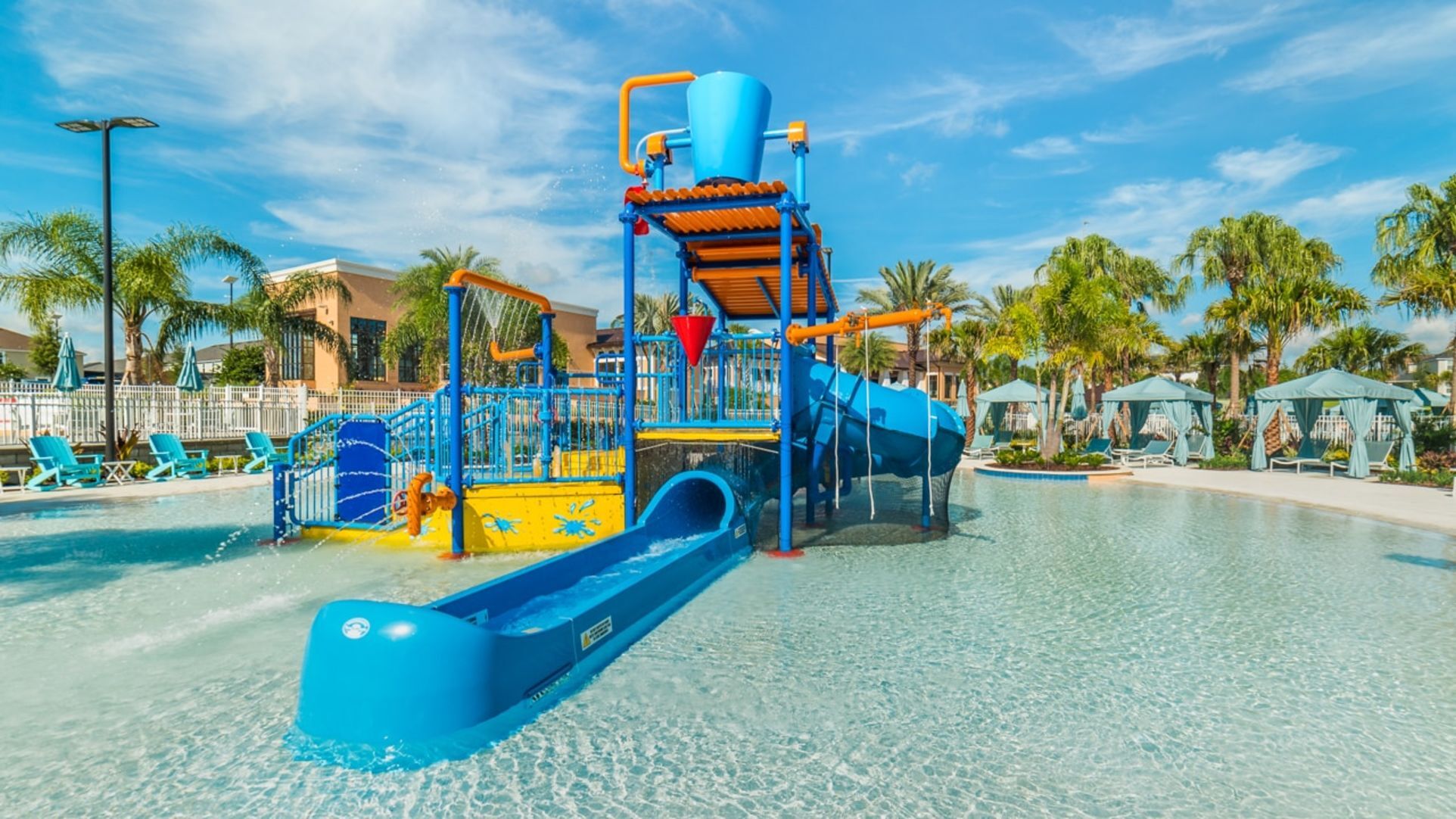 A bright blue and yellow children’s splash pad and water play structure with slides and a tipping bucket at Solara Resort, surrounded by cabanas and a zero-entry pool.