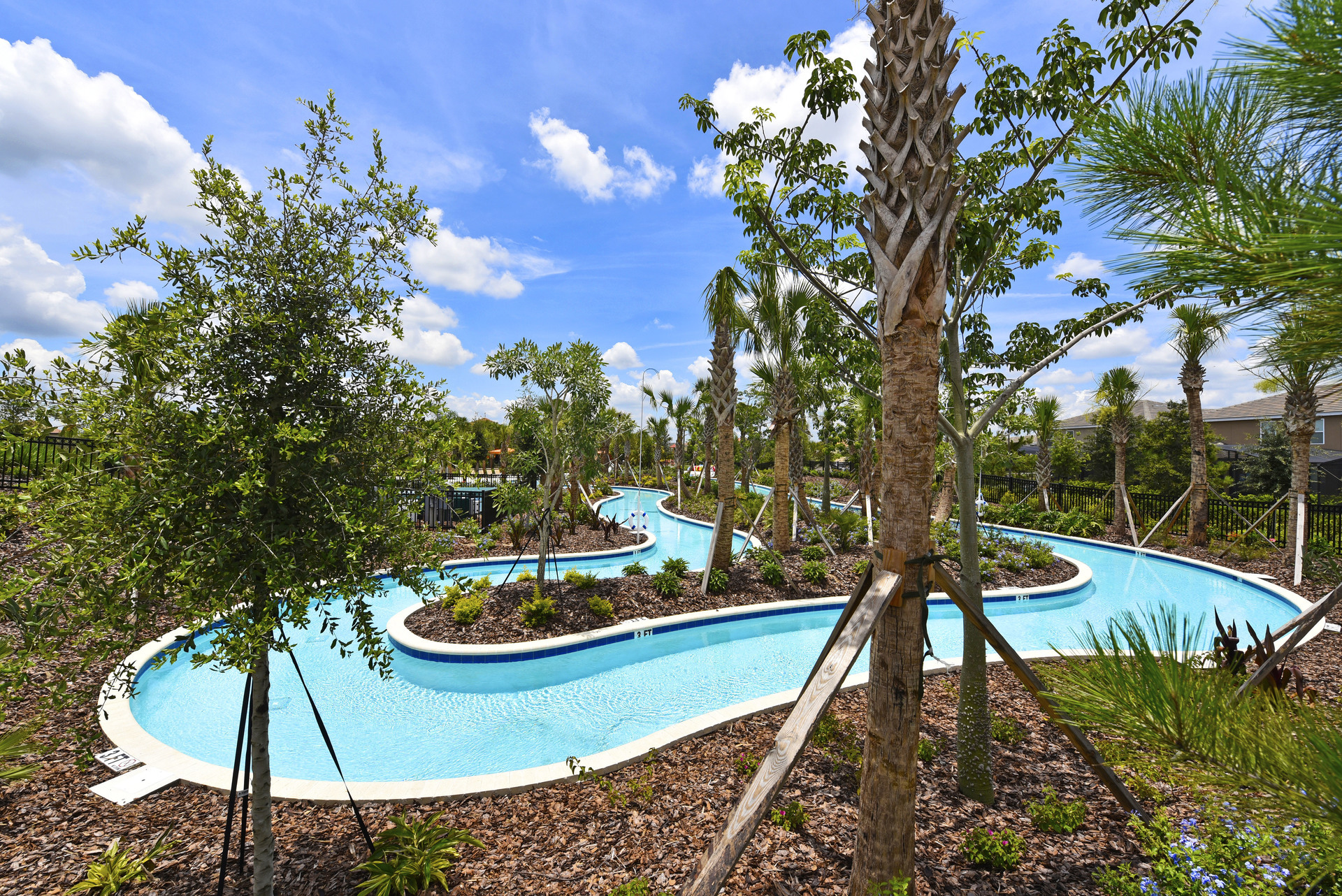 A bright blue, winding lazy river pool at Solterra Resort in Orlando, surrounded by young palm trees, lush landscaping, and a clear blue sky with clouds.