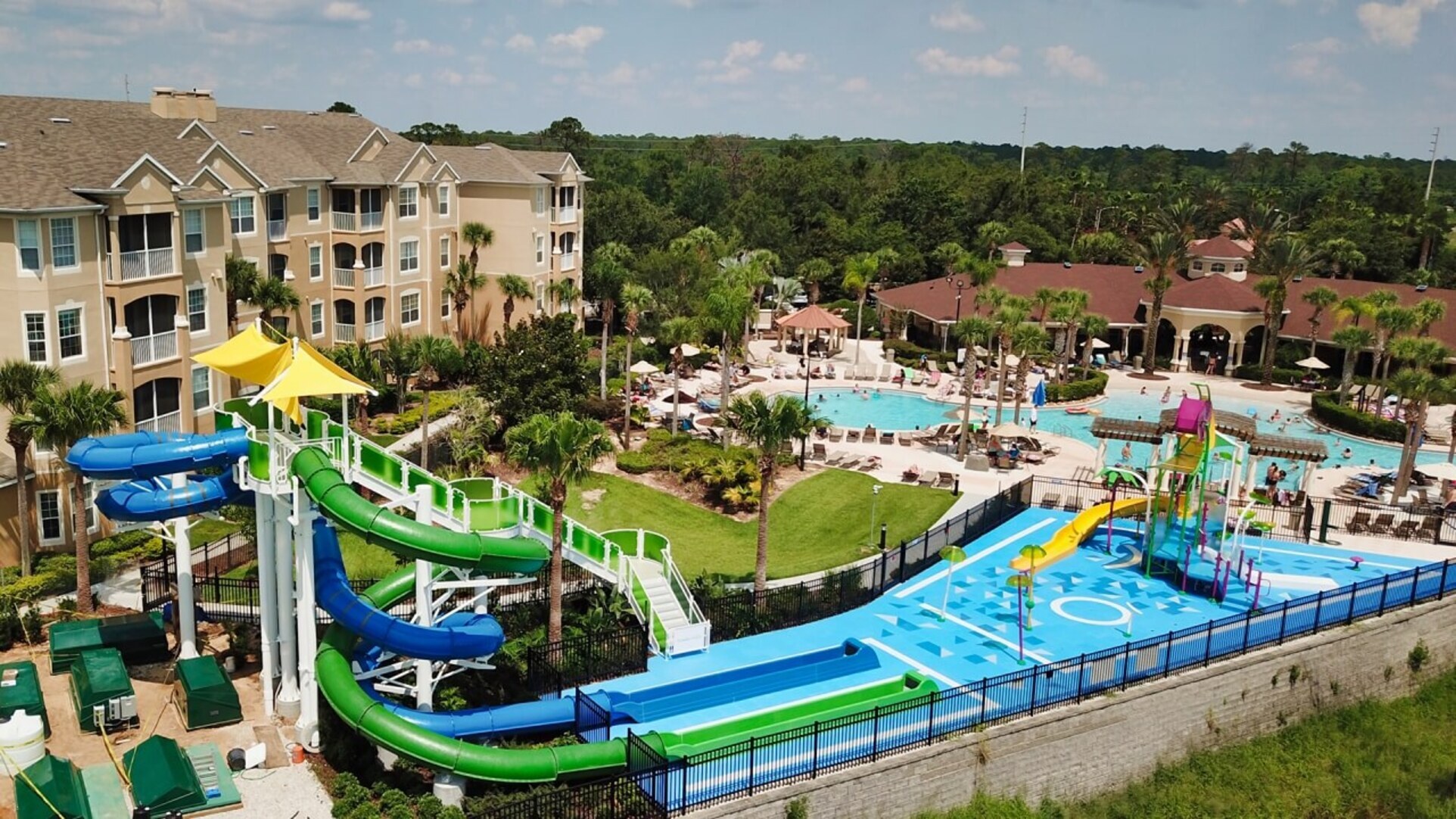 An aerial view of the Windsor Hills Resort water park in Orlando, featuring large green and blue winding water slides, a children’s splash pad area, and a large lagoon-style pool surrounded by palm trees and resort buildings.